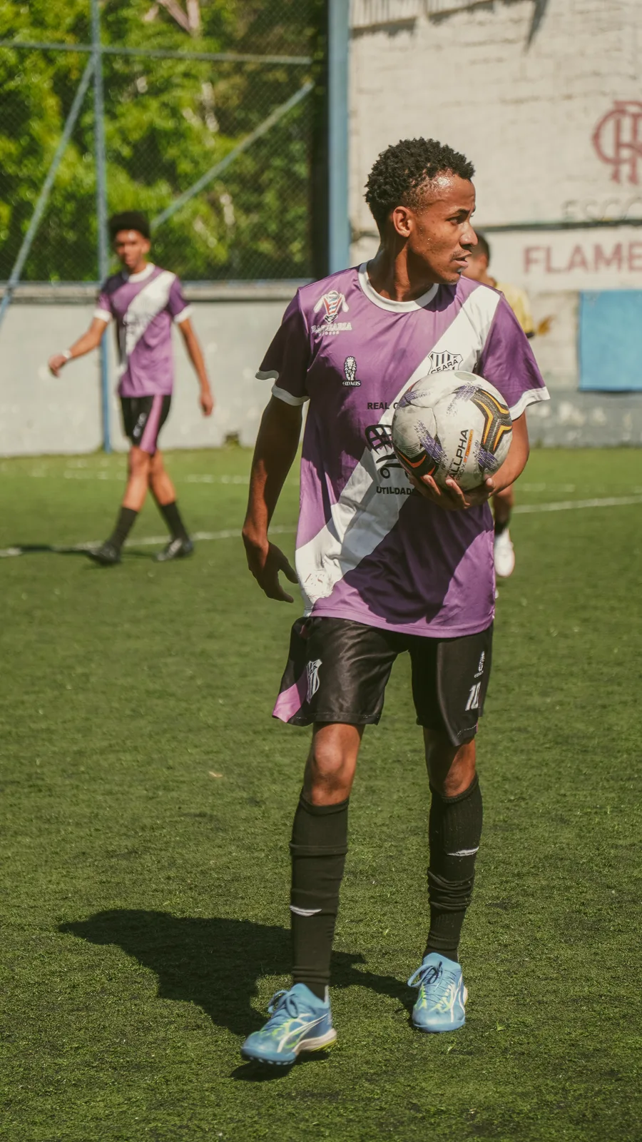 Jogador de uniforme roxo segura a bola ao lado de um companheiro de time