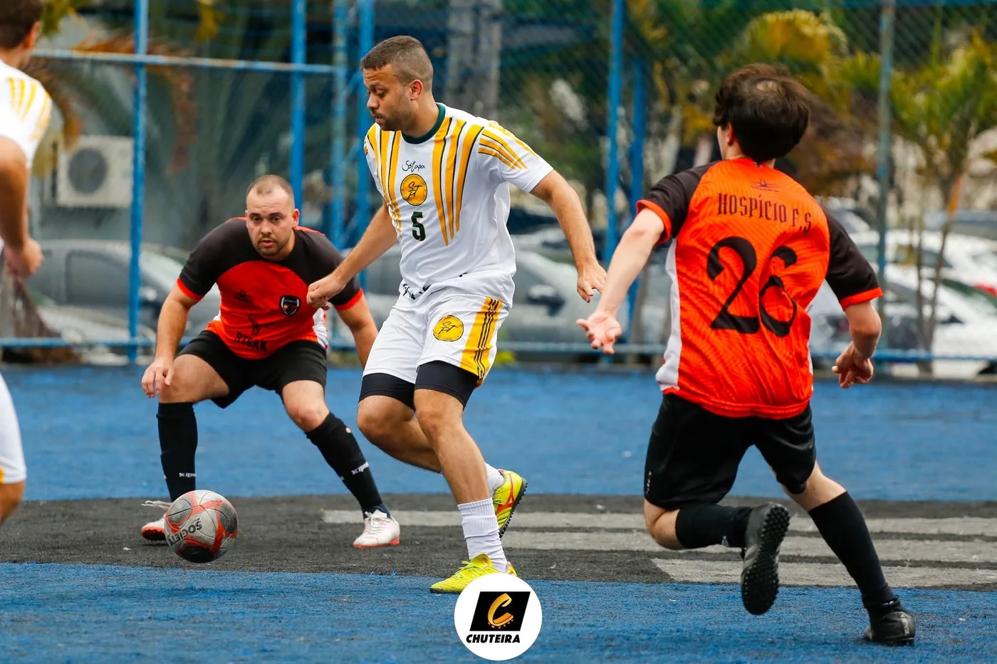 Jogador de branco e amarelo conduz a bola entre adversários de laranja em campo sintético