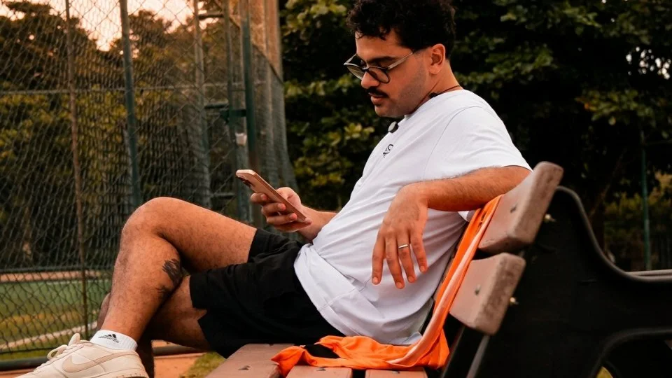 Pessoa com camiseta branca do NaTrave consulta o celular sentada em um banco ao lado do campo