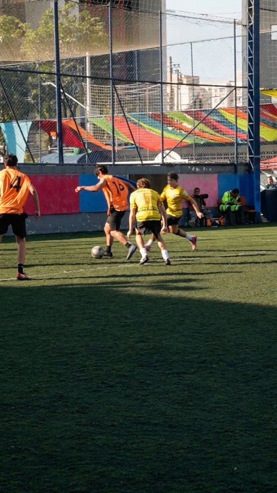 Jogadores de coletes amarelos e laranjas disputam a bola em uma quadra de grama sintética